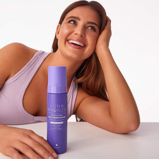 A smiling woman in a light lavender tank top leans on a white surface next to a purple bottle of Luna Bronze Ultra-Luxe Hair Tanning Mousse.